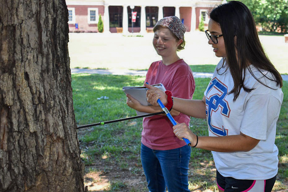 One college student bores a hole into a tree with a tree corer while another student holds a notebook and looks on. | Adrianna Cody and Sharmila Samuel | Presbyterian College | Clinton SC