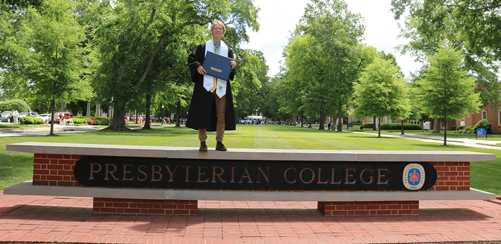 Graduate student with his diploma in front of Presbyterian College sign
