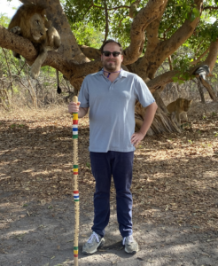 Dr. Ben Bailey stands with a hiking stick with a lion in a tree behind him.