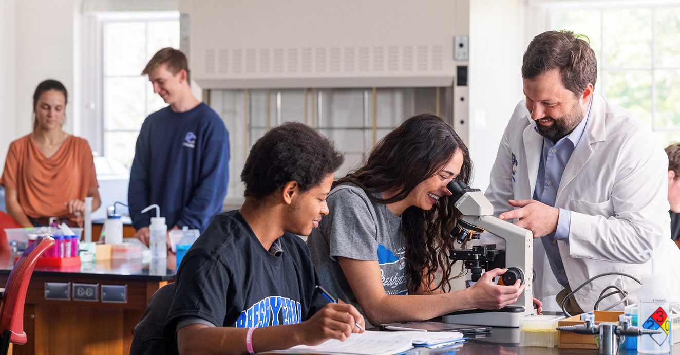 Two students are doing research, one is writing, while another is looking into a microscope, Dr. Austin Shull is to the right of them and is helping one of the students as they look into the microscope