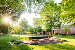 A picture of the Presbyterian College sign with the morning sun peaking through the trees.